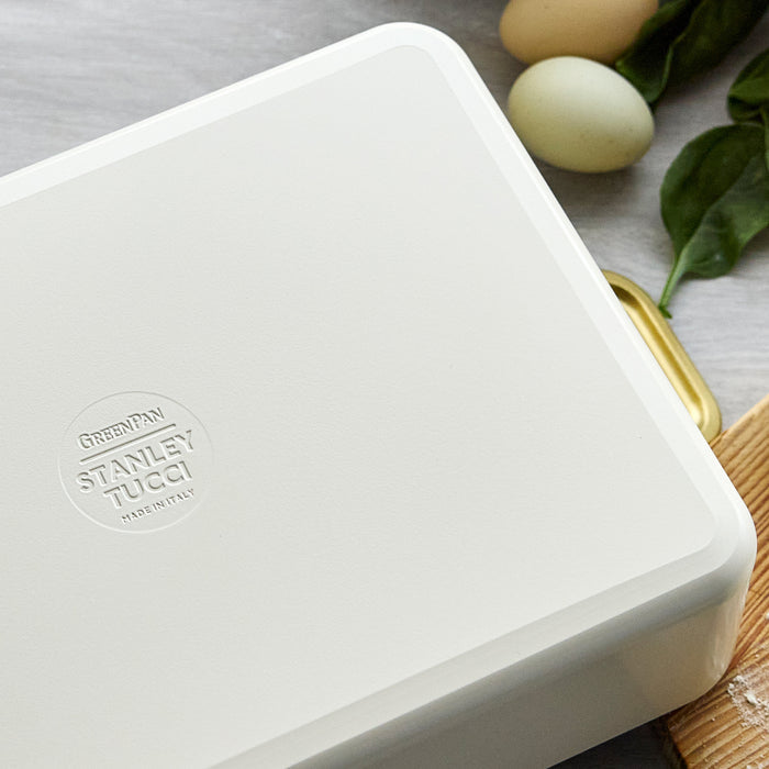 White ceramic lasagna pan on a table with brown eggs, fresh greens, and a wooden cutting board.