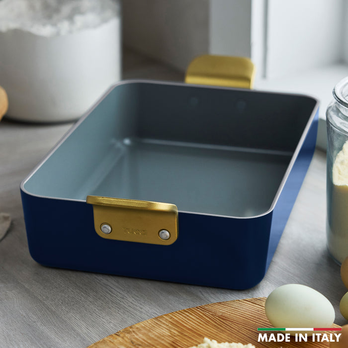 Ceramic nonstick lasagna pan in blue with gold handles, on a kitchen counter with baking items.