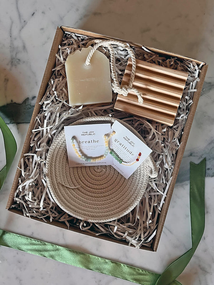 Gift box featuring handmade soap with cord, wooden soap dish, 'Breathe' bracelet, and 'Gratitude' bracelet, on green ribbon.