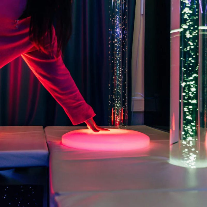 Hand reaching down to the glowing red TFH Illuminated Floor Splot beside bubble tubes, an interactive sensory light for multi-sensory rooms