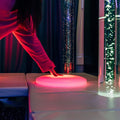 Hand reaching down to the glowing red TFH Illuminated Floor Splot beside bubble tubes, an interactive sensory light for multi-sensory rooms