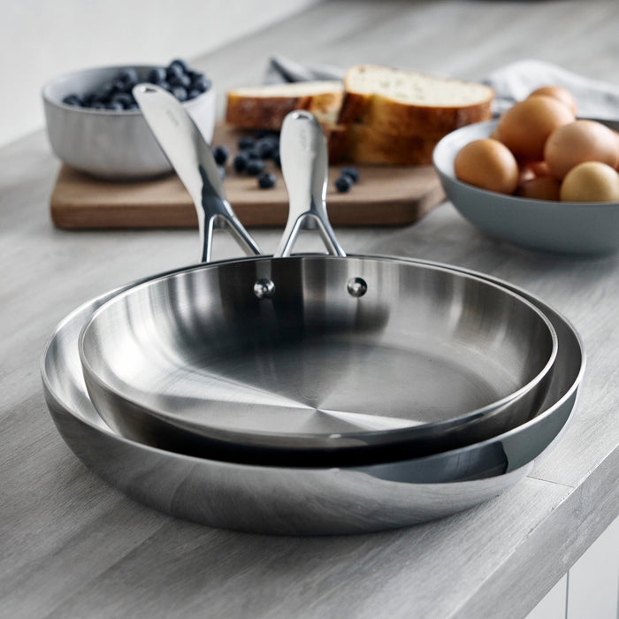 Two stainless steel frying pans stacked on a kitchen counter with eggs, bread, and blueberries nearby.