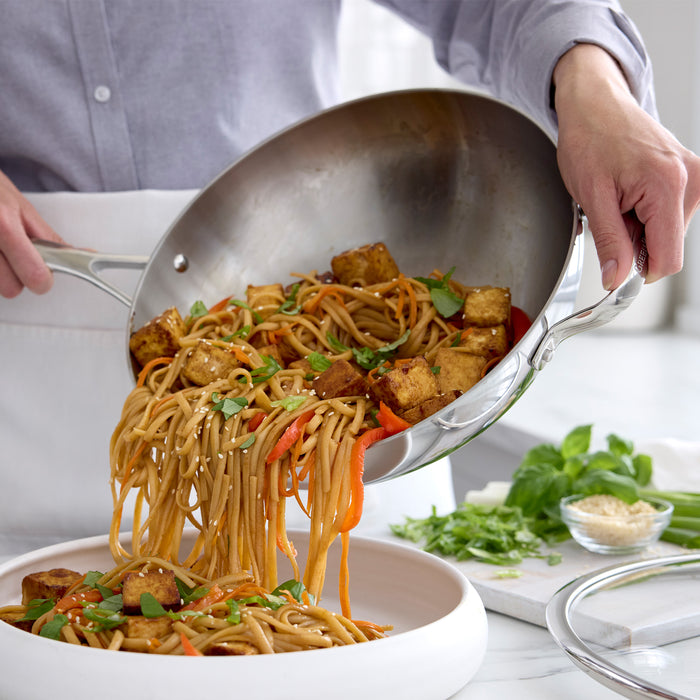 Stir-fried noodles and veggies are poured from a stainless steel wok into a white bowl.