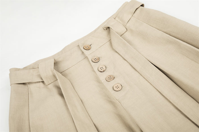 Close-up of beige trousers showing button fly, pleats, and waist tie belt