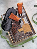 A curated gift set in a cardboard box with white crinkle paper, featuring a black satin sleep mask, two orange tubes from The Riches, a brown leopard-print pouch, a tan leather luggage tag, and a matching leather card holder, arranged on a marble surface with green ribbon and leaf accents.